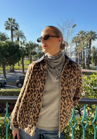 Femme portant une veste à imprimé léopard, une écharpe grise, des lunettes de soleil et un chignon, debout en extérieur avec des palmiers et des voitures en arrière-plan.