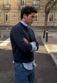 Νεαρός άνδρας με σταυρωμένα χέρια, φορώντας σκοτεινό πουλόβερ πάνω από μπλε πουκάμισο και τζιν, στέκεται σε δρόμο της πόλης κοντά σε πεζοδρόμιο και κτίρια.