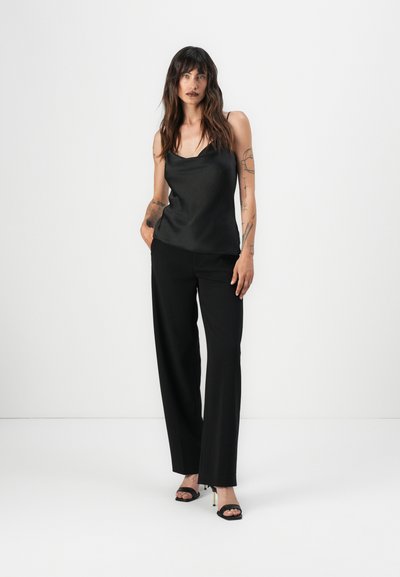 Camisón de satén negro con escote drapeado, combinado con pantalones negros de talle alto. Presenta un corte holgado y líneas verticales. Tacones de aguja negros.