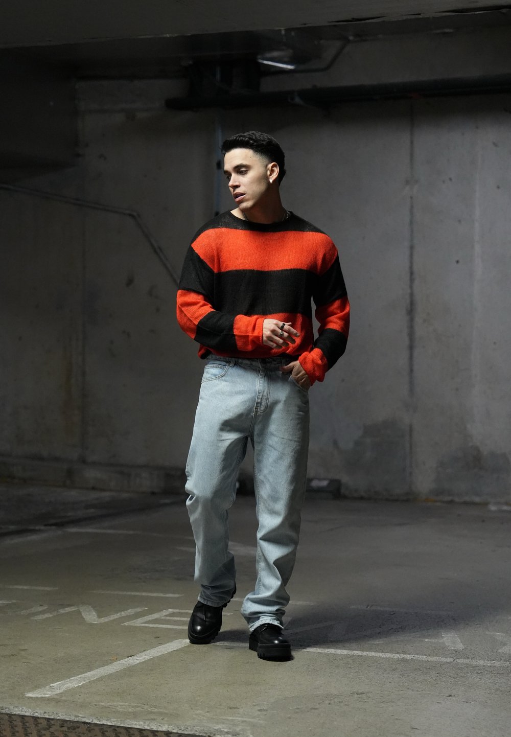 Μαύρη και πορτοκαλί ριγέ ζακέτα, ανοιχτού γαλάζιου χρώματος τζιν παντελόνι και μαύρα παπούτσια. Φόντο είναι μια τσιμεντένια περιοχή με ουδέτερους τόνους και ελάχιστες λεπτομέρειες.