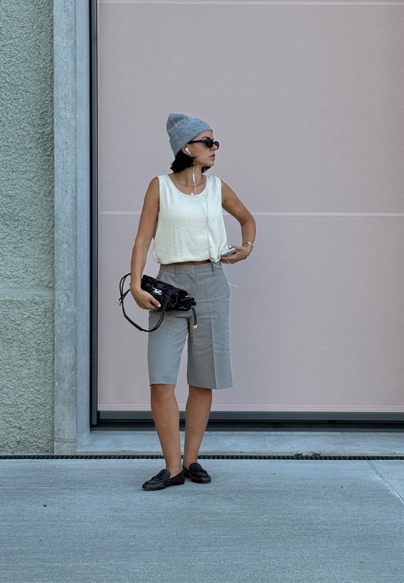 Leggero top oversize grigio chiaro, pantaloni corti grigi su misura, mocassini neri, berretto grigio, tiene una borsa scura, con occhiali da sole e cuffie.