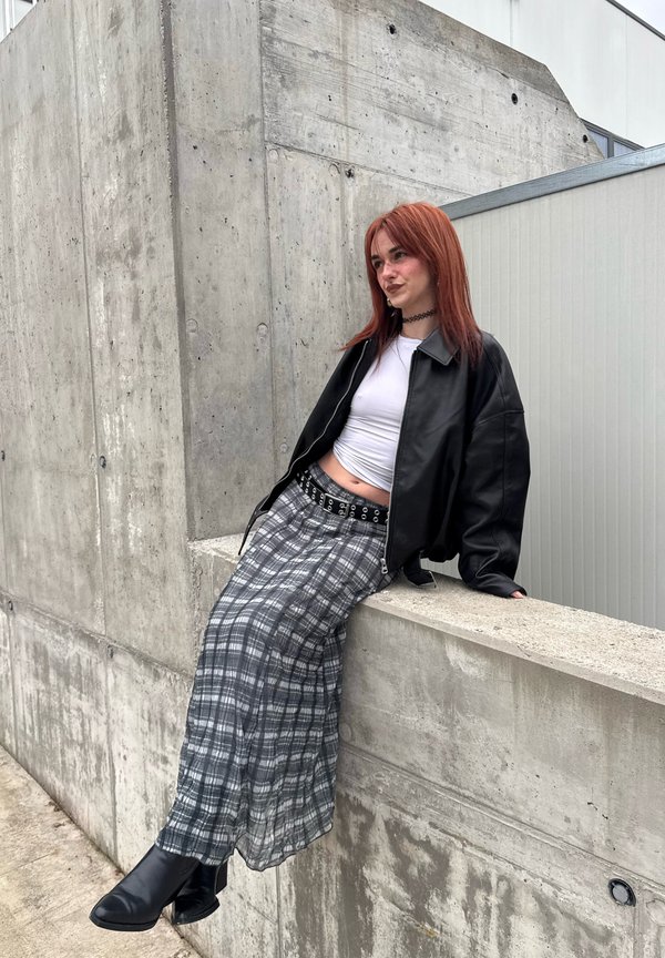 Tânăr cu păr roșcat, purtând o jachetă neagră din piele, top alb scurt, fustă în carouri și cizme, așezat pe o margine de beton în aer liber.