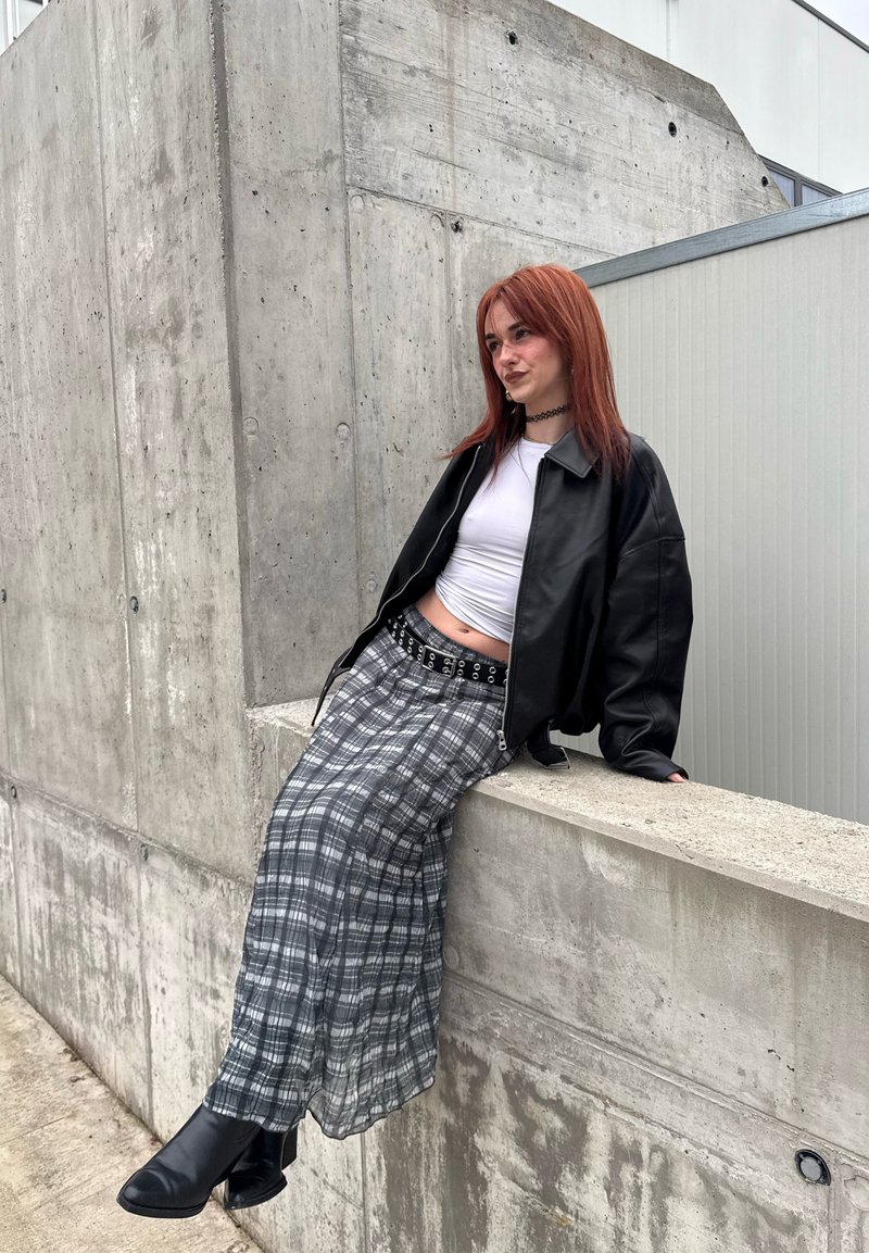 Młoda osoba z rudymi włosami, ubrana w czarną skórzaną kurtkę, biały krótki top, spódnicę w kratę i buty, siedząca na betonowym brzegu na zewnątrz.