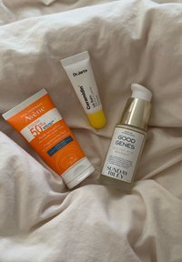 Drei Hautpflegeprodukte auf einem hellen Stoff: eine orange Sonnencremetube mit weißen Akzenten, ein gelber Lippenbalsam und eine Flasche mit Glykoläurebehandlung und silbernem Deckel.