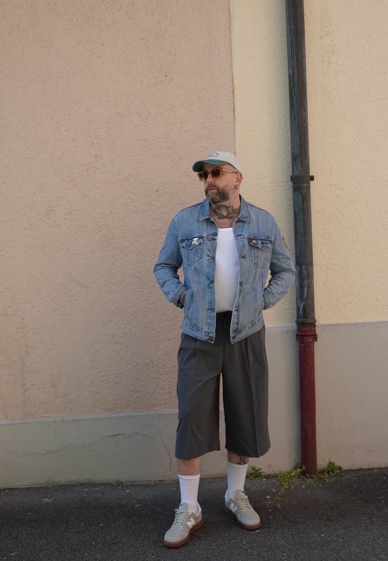 Homme avec barbe et tatouages portant une veste en denim, une casquette, des lunettes de soleil, un short long à fines rayures, des chaussettes blanches et des baskets, debout contre un mur.