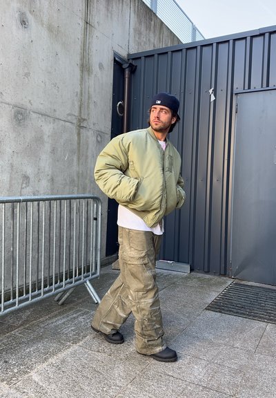 Cazadora bomber verde oliva, camiseta blanca, pantalones cargo caqui y zapatillas negras, situados frente a paredes de hormigón y vallas metálicas.