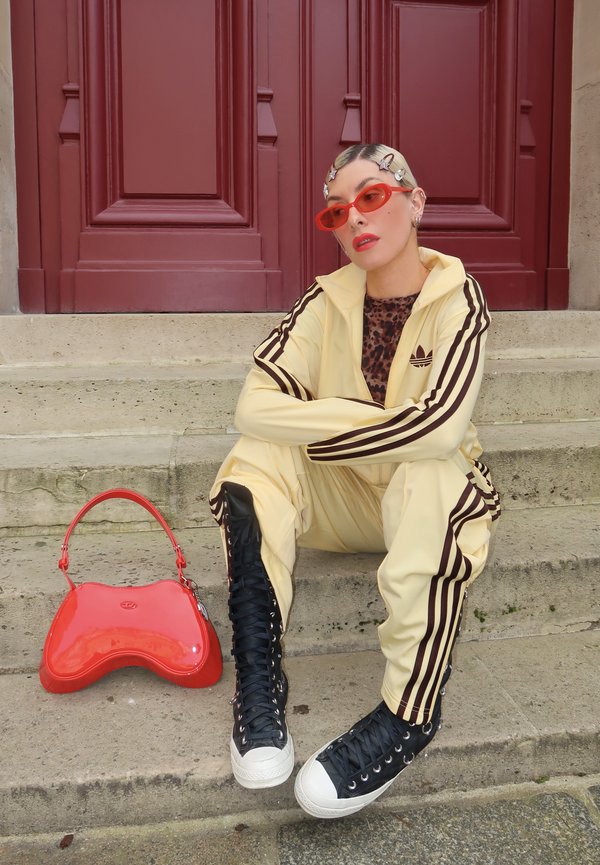 Personne aux cheveux blonds avec des pinces, portant un survêtement jaune Adidas, un haut imprimé léopard, des lunettes de soleil rouges et des baskets montantes noires, assise près d'une porte rouge.