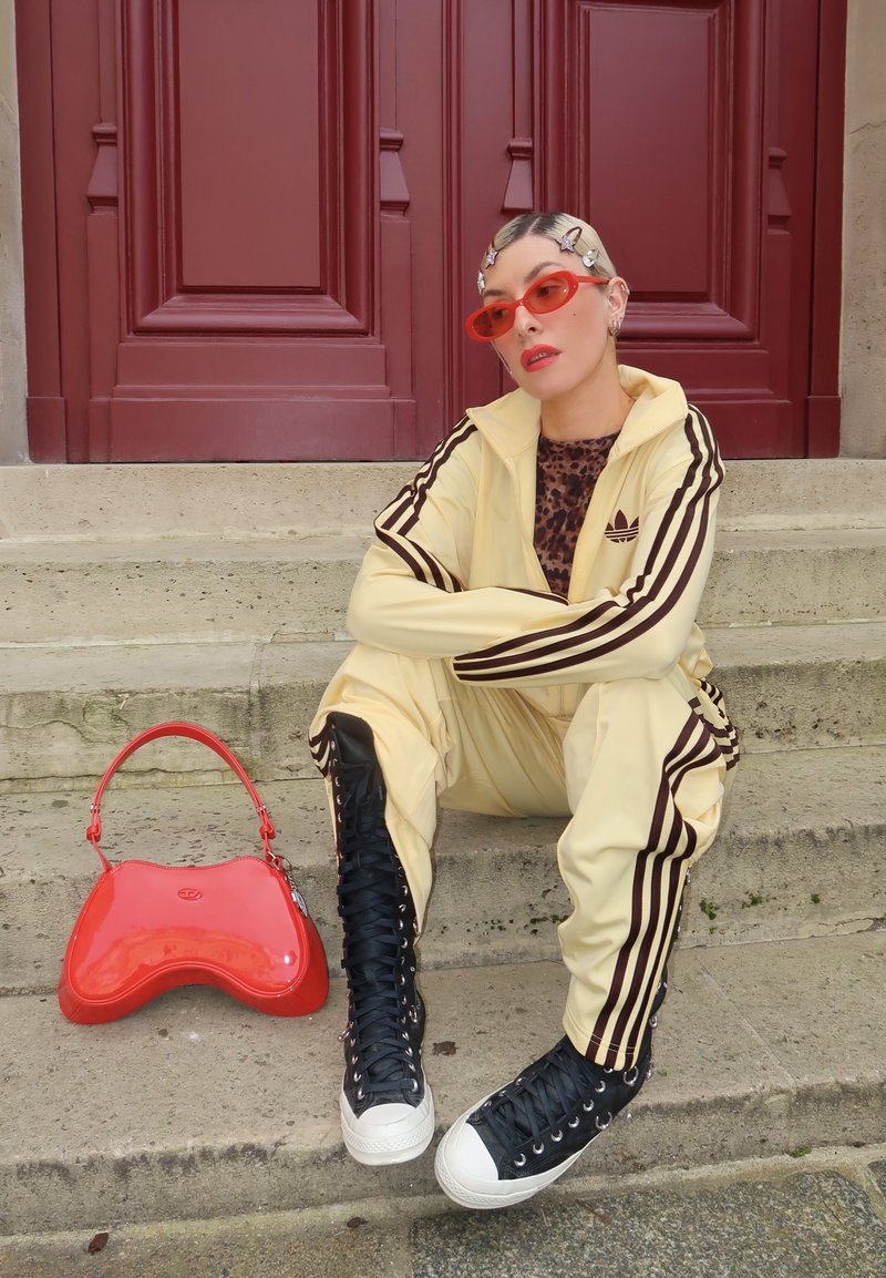 Osoba se světlými vlasy a sponkami má na sobě žlutou teplákovou soupravu Adidas, leopardí top, červené sluneční brýle a černé kotníkové tenisky, sedí u červených dveří.