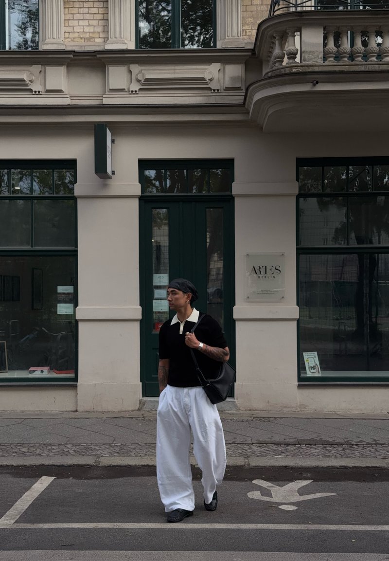 Homme avec une chemise noire et un pantalon blanc portant un sac noir, marchant dans la rue devant un bâtiment avec des portes et fenêtres vertes.