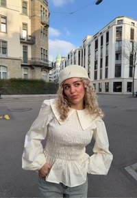 Valkoinen, rypytetty paita, jossa on suuri kaulus ja puffihihat, yhdistettynä vaaleisiin farkkuihin. Lookkia täydentää kreeminvärinen Beret, jossa on brodeerattu logo.