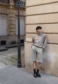 Lysegrå oversized genser, plisserte lyse beige shorts, svarte sokker og svarte loafers. Står ved en teksturert steinvegg og en lyktestolpe.