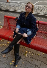 Denim φόρεμα με ανοιχτόχρωμες λεπτομέρειες από τζιν, φορεμένο πάνω από μια σκούρα τζιν πουκάμισο. Μαύρες μπότες μέχρι το γόνατο με μυτερές μύτες. Καθισμένη σε μία κόκκινη τρυπητή παγκάκι.