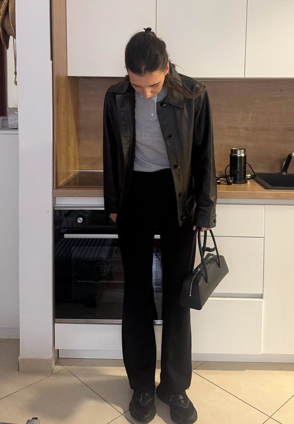 Une femme baisse les yeux vers sa tenue noire et son sac à main dans une cuisine moderne.
