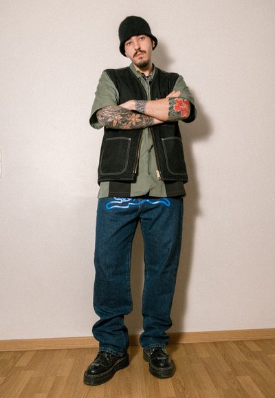 Chaleco negro sin mangas sobre una camisa verde, jeans bordados azules y zapatos negros. Tatuajes notables en los brazos y un gorro negro.