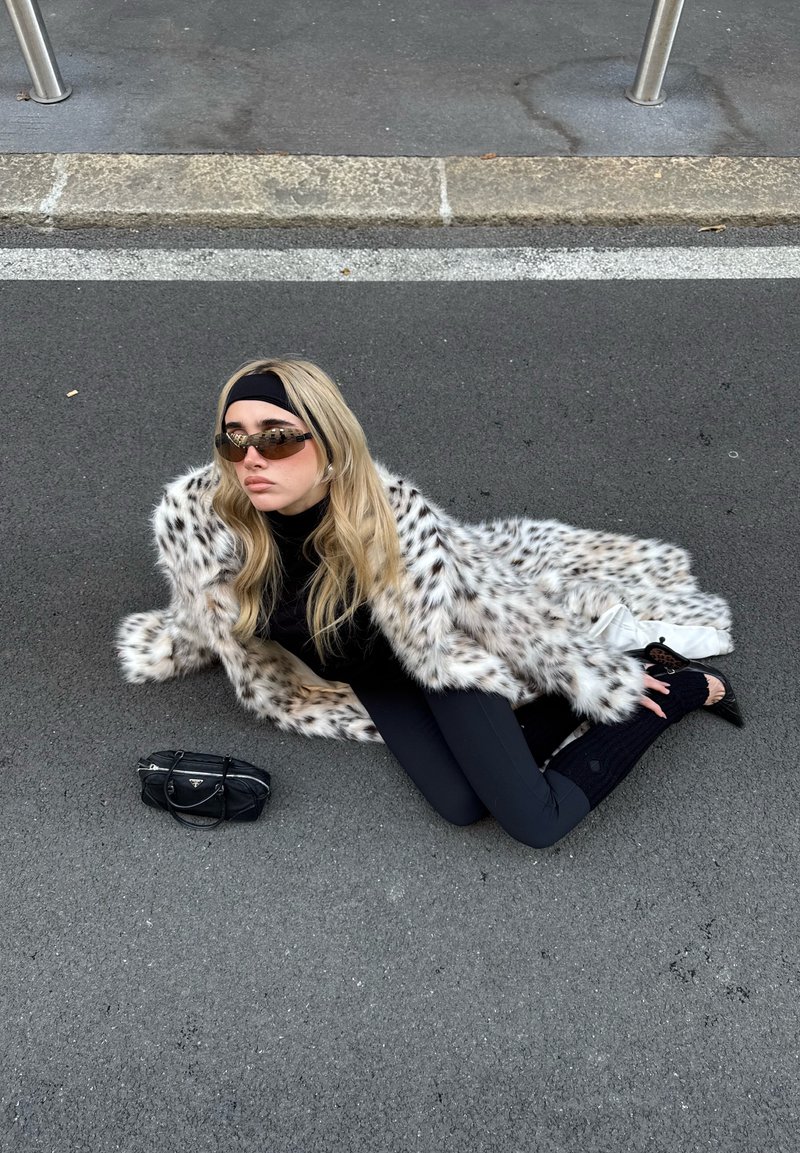 Manteau en fausse fourrure à motif léopard, col roulé noir, leggings et chaussures à talons avec découpes. Accessoires comprenant un petit sac à main noir et des lunettes de soleil.