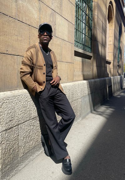 Cardigan de patrón marrón y negro sobre una camiseta negra, combinado con pantalones negros y zapatos negros gruesos, de pie contra una pared de piedra.