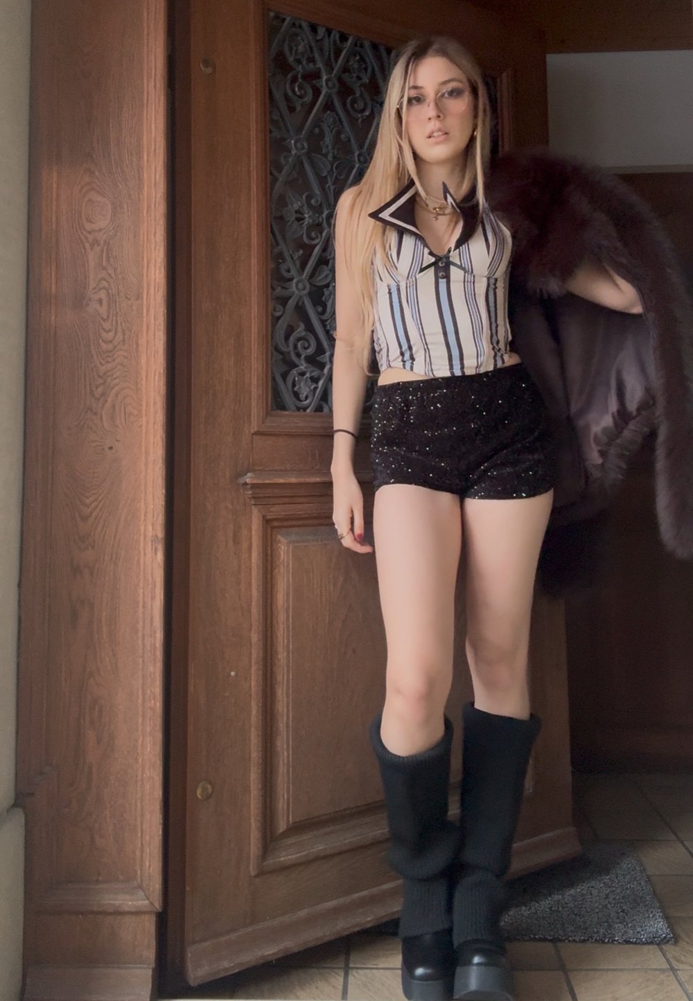 Jeune femme posant devant une porte en bois, vêtue d'un haut rayé sans manches, d'un short noir à sequins, de longues bottes noires, et tenant un manteau de fourrure sombre.