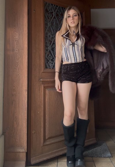 Joven de pie frente a una puerta de madera, vestida con una camiseta sin mangas a rayas, pantalones cortos negros con lentejuelas, botas altas negras y sosteniendo un abrigo de piel oscuro.