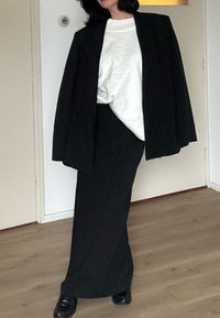 Personne portant une veste de costume noire à fines rayures sur les épaules, une jupe longue assortie, un pull blanc et des chaussures noires, debout sur un sol en bois à l'intérieur.