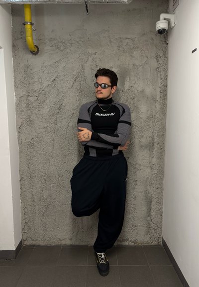 Camisa ajustada de manga larga en negro y gris con rayas, pantalones negros holgados y zapatillas negras. Hombre apoyado contra una pared de concreto texturizada.