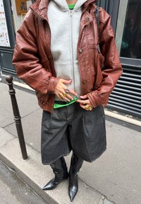 Person iført brun skinnjakke over grå hettegenser og grønn skjorte, svarte shorts med vide ben, svarte spisse støvler, og flere ringer på fingrene.