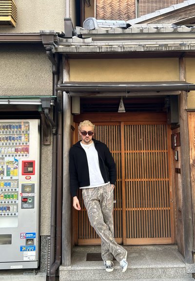 Cárdigan de punto negro sobre una camiseta blanca, pantalones con patrón de camuflaje y zapatillas deportivas grises. De pie junto a una puerta de madera y una máquina expendedora.