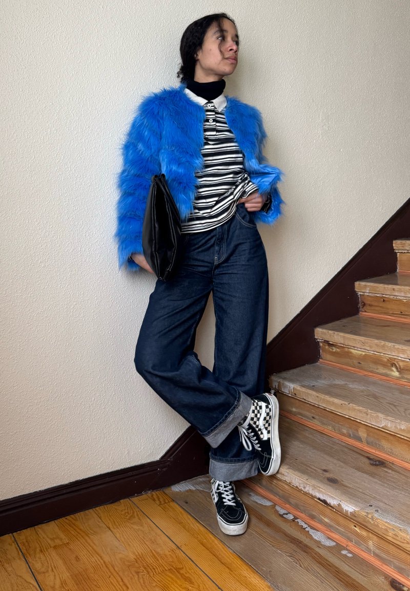 Personne appuyée contre un mur près d'un escalier en bois, portant une veste en fausse fourrure bleue, une chemise rayée, un jean large, des baskets à carreaux, tenant une pochette noire.