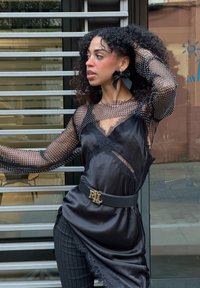 Sort mesh langærmet top med sølvnitter, lagdelt over en sort satin kjole. Har blondekant og et sort bælte med gyldent emblem.