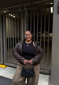 Veste oversize en cuir marron, débardeur noir, pantalon large beige, tenant un petit sac en nylon noir avec un logo. Fond gris avec des portes.