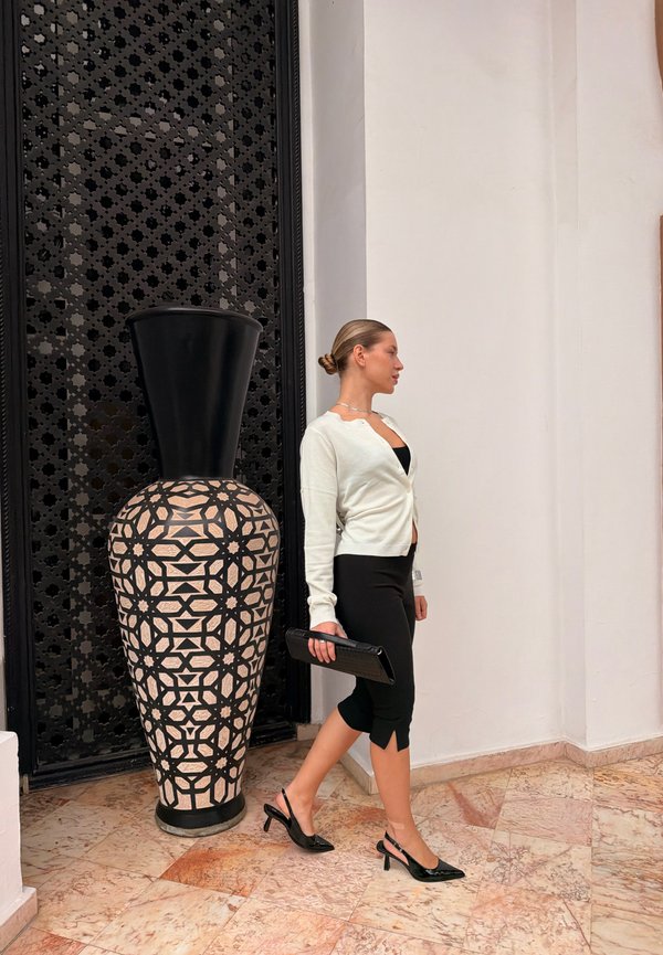 Frau in schwarzem Rock und weißem Cardigan, die eine Clutch hält und neben einer großen schwarz-beigen dekorativen Vase steht.