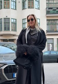 Femme portant un manteau noir à poignets en fourrure, une écharpe grise et des lunettes de soleil, tenant un sac à main noir dans une rue en ville avec des voitures et des immeubles.