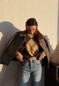 Femme portant un manteau à carreaux par-dessus un pull à motif losanges et un jean bleu, ajustant son pantalon dans une lumière intérieure chaude.