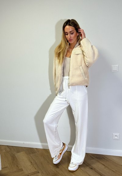 Chaqueta de forro polar beige con textura y cierre de cremallera, combinada con pantalones blancos y zapatillas con acentos en naranja. Suelo de madera, fondo sencillo.