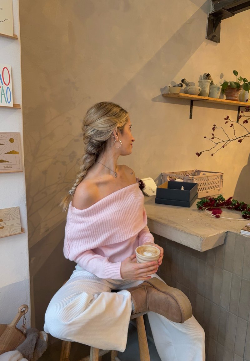 Light pink off-shoulder ribbed sweater, cream wide-leg pants, brown ankle boots, holding a coffee cup, seated on a wooden stool.