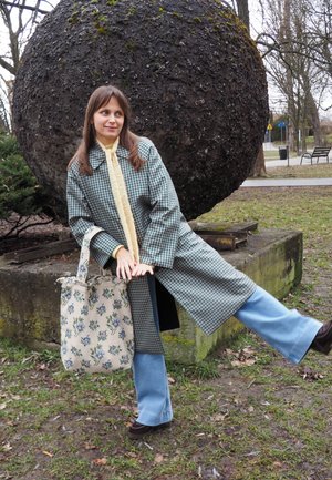 Joven mujer con abrigo a cuadros y vaqueros azules posando con una pierna levantada junto a una gran esfera de piedra texturizada en un parque.