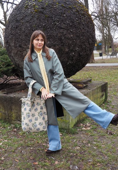 Joven mujer con abrigo a cuadros y vaqueros azules posando con una pierna levantada junto a una gran esfera de piedra texturizada en un parque.