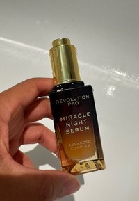 Flacon de sérum en verre noir avec un piston doré, étiqueté "Revolution Pro Miracle Night Serum", présentant un dégradé allant d'un ambre foncé à transparent.
