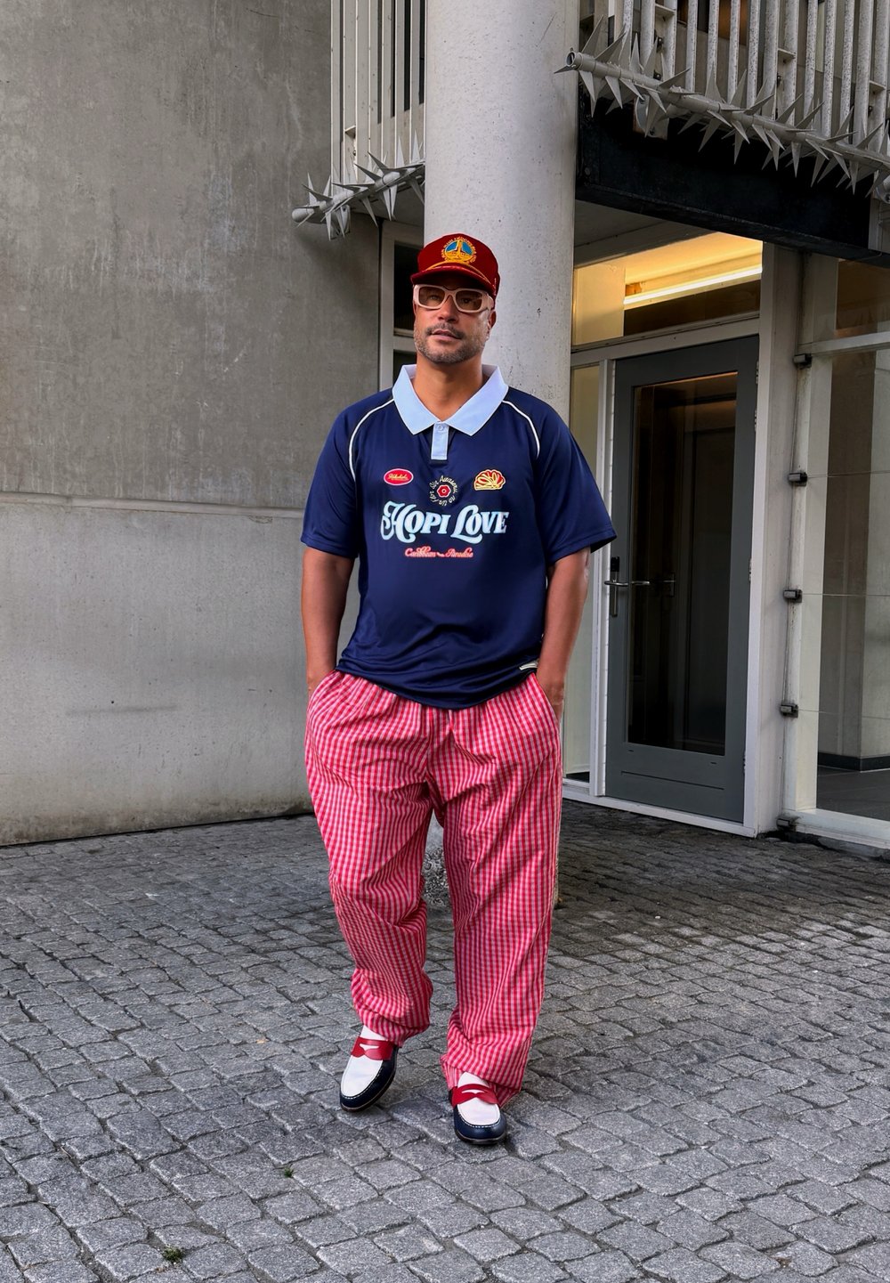 Polo navy con colletto bianco e testo "HOPI LOVE"; pantaloni larghi a scacchi rossi e bianchi; scarpe nero e bianche con dettagli rossi.