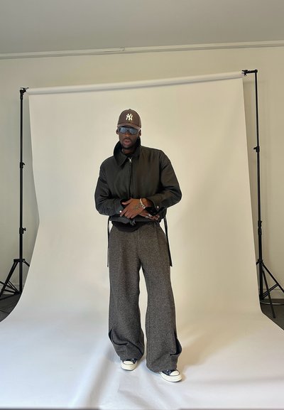 Hombre con gorra marrón, gafas de sol, chaqueta negra, pantalones anchos grises y zapatillas posando frente a un fondo blanco para fotografía.