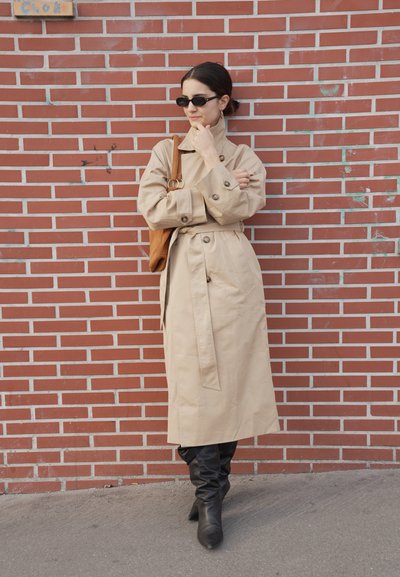 Mujer con gabardina beige y botas negras, de pie contra una pared de ladrillos rojos, con gafas de sol negras y llevando un bolso marrón de hombro.
