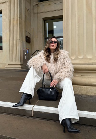 Mujer con un abrigo beige de peluche, pantalones blancos, botas negras de tacón y gafas de sol, sentada en unos escalones de piedra sosteniendo un bolso negro junto a una columna.