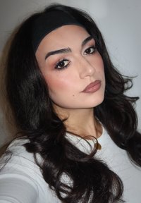 Portrait d'une personne aux cheveux longs, ondulés et foncés, portant un bandeau noir et un haut blanc. Le maquillage présente des tons naturels et des sourcils définis.