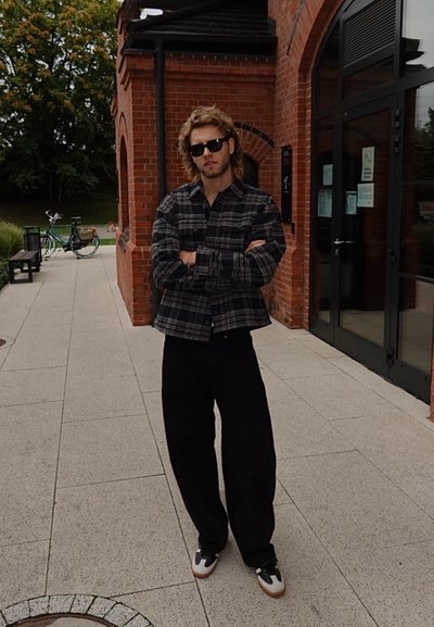 Camisa de cuadros gris de gran tamaño, pantalones anchos negros y zapatillas gris y blancas. Hombre con cabello rizado y gafas de sol se encuentra de pie frente a un fondo de ladrillo.