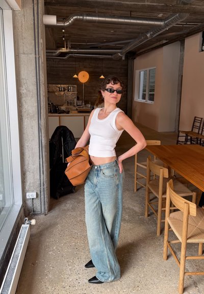 Mujer con camiseta sin mangas blanca y jeans anchos sosteniendo una bolsa marrón, de pie junto a sillas y una mesa de madera en una habitación de estilo industrial moderno.