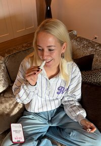 Tube de baume à lèvres blanc tenu près de la bouche, porté avec un pull gris clair à rayures et un pantalon bleu ample, assis sur un canapé marron texturé.