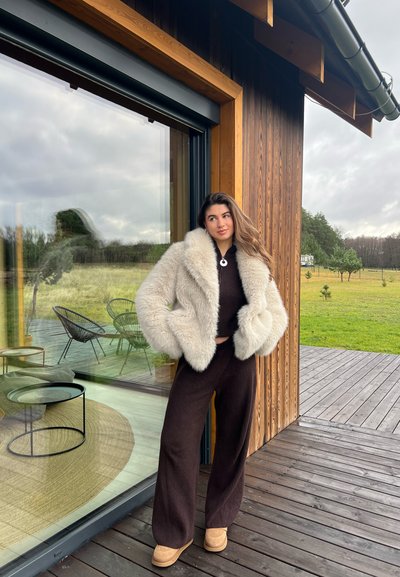 Chaqueta de piel sintética en color crema con amplio cuello, top marrón de canalé y pantalones de pierna ancha a juego, combinados con zapatos de plataforma en color beige. Fondo de madera.