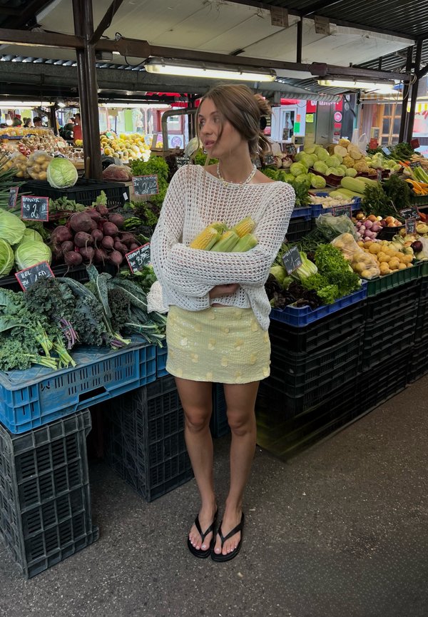 Donna con maglione bianco e gonna gialla che tiene del mais, in piedi in una bancarella di mercato circondata da verdure fresche.