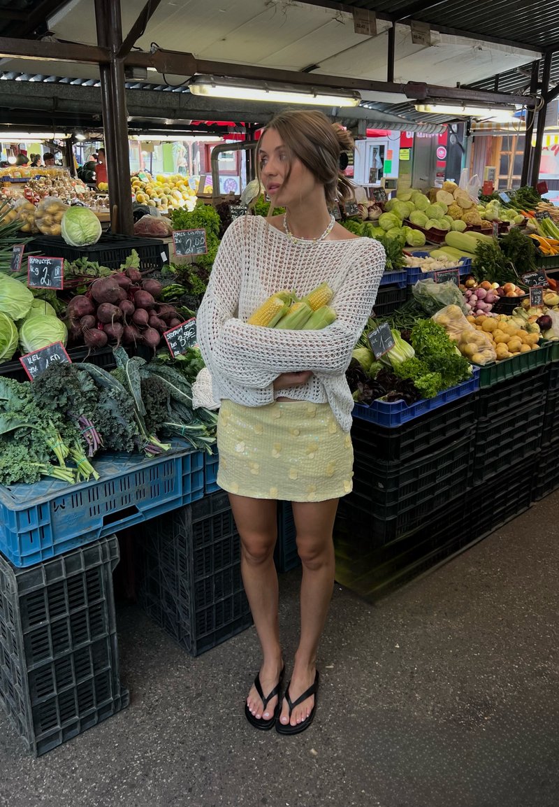 Woman in a white sweater and yellow skirt holding corn stands in a market stall surrounded by fresh vegetables.