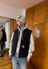 Άνδρας που φοράει μπεζ σκουφάκι, γκρι φούτερ, μαύρο γιλέκο και τζιν, στέκεται σε εσωτερικό χώρο κοντά σε ξύλινα ντουλάπια και έναν καθρέφτη που αντανακλά το πλάι του προφίλ.