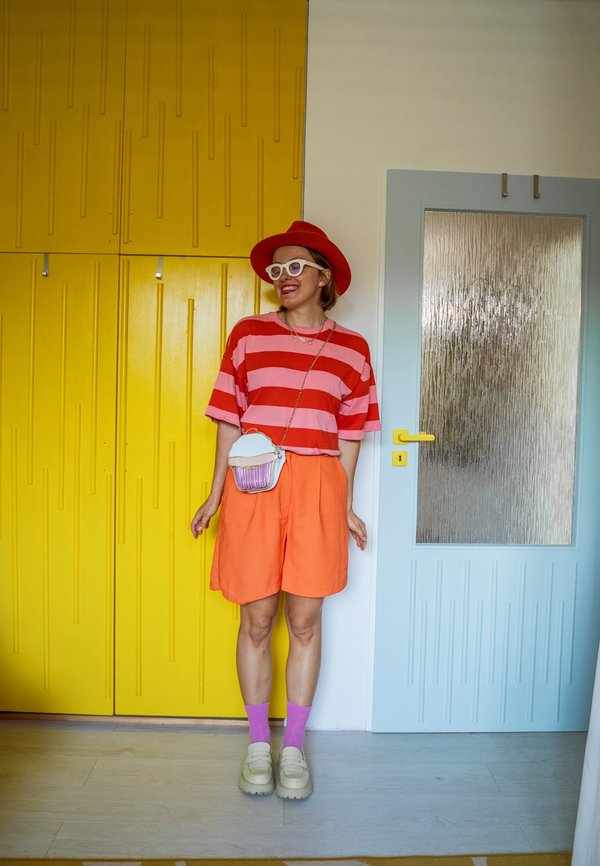 Kvinde iført rød og pinkstribet skjorte, orange shorts, rød hat, hvide briller, pink sokker og cremefarvede sko, står ved gule skabe og en blå dør.